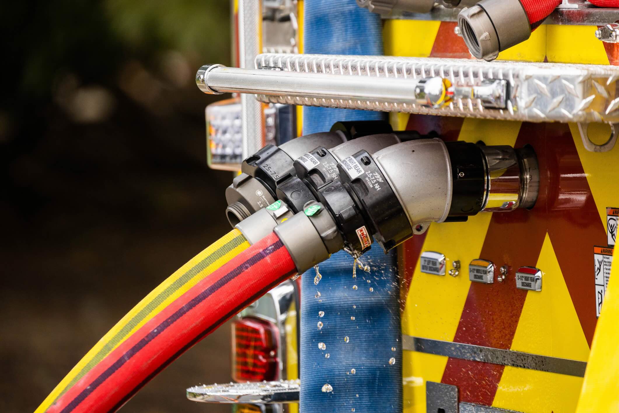 A close-up, dynamic view of a high-pressure water hose extending from a fire truck and actively being used by firefighters to spray water onto a fire scene