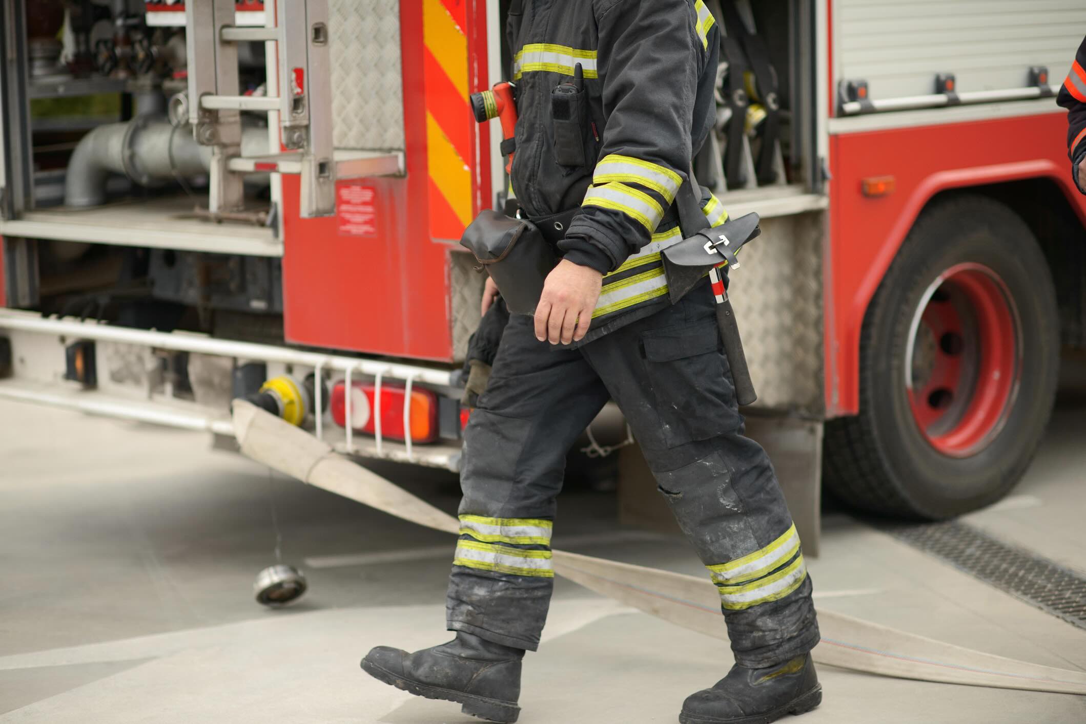 A firefighter in full turnout gear walks beside an active fire truck with a hose on the ground, showing emergency readiness with visible tools, safety stripes, and open compartments.